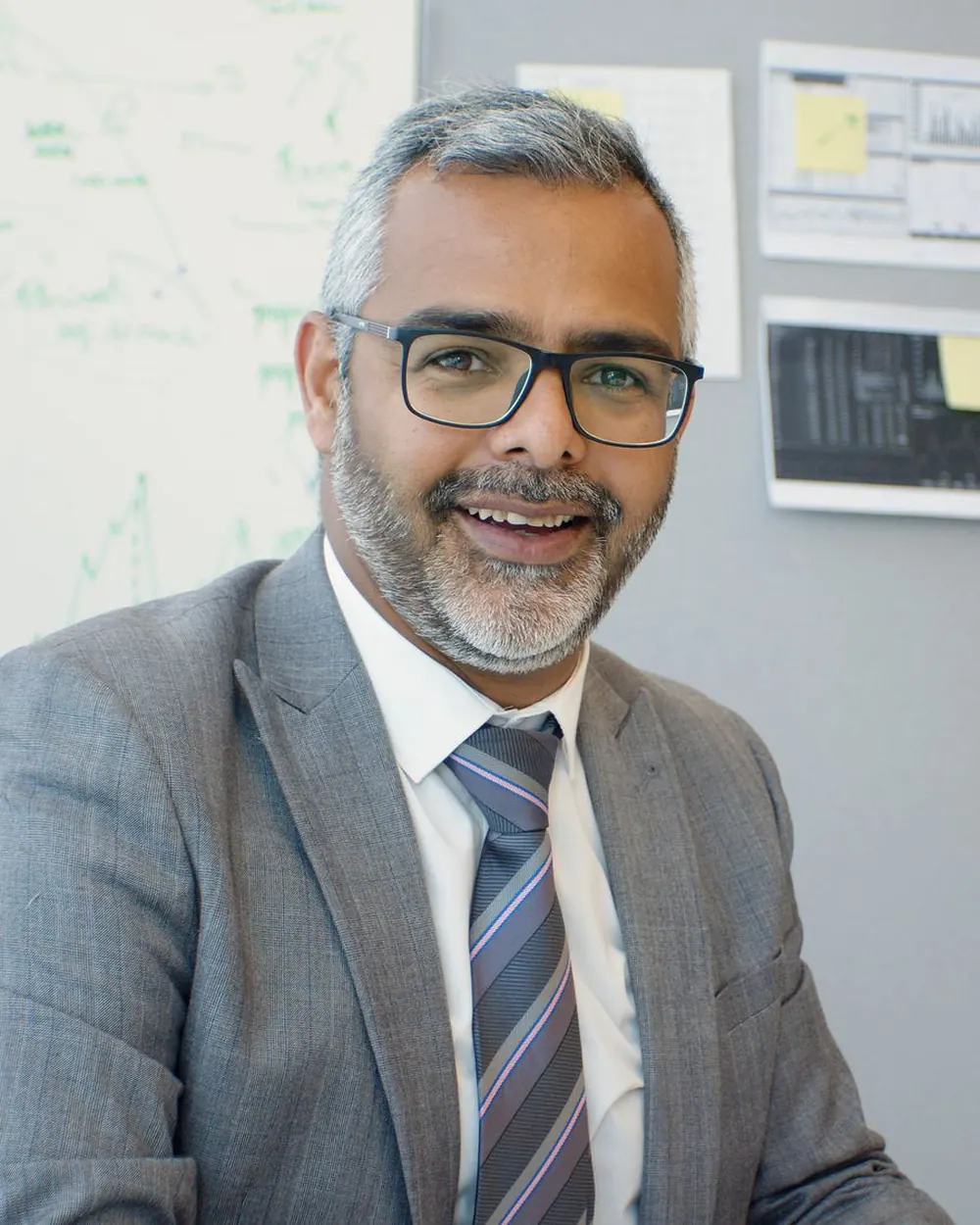 A smiling senior professional in a grey suit at his office desk